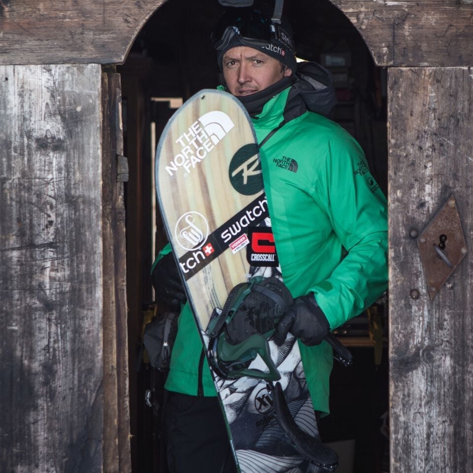 Xavier De Le Rue holding snowboard, ready to go, exiting wooden shed workshop